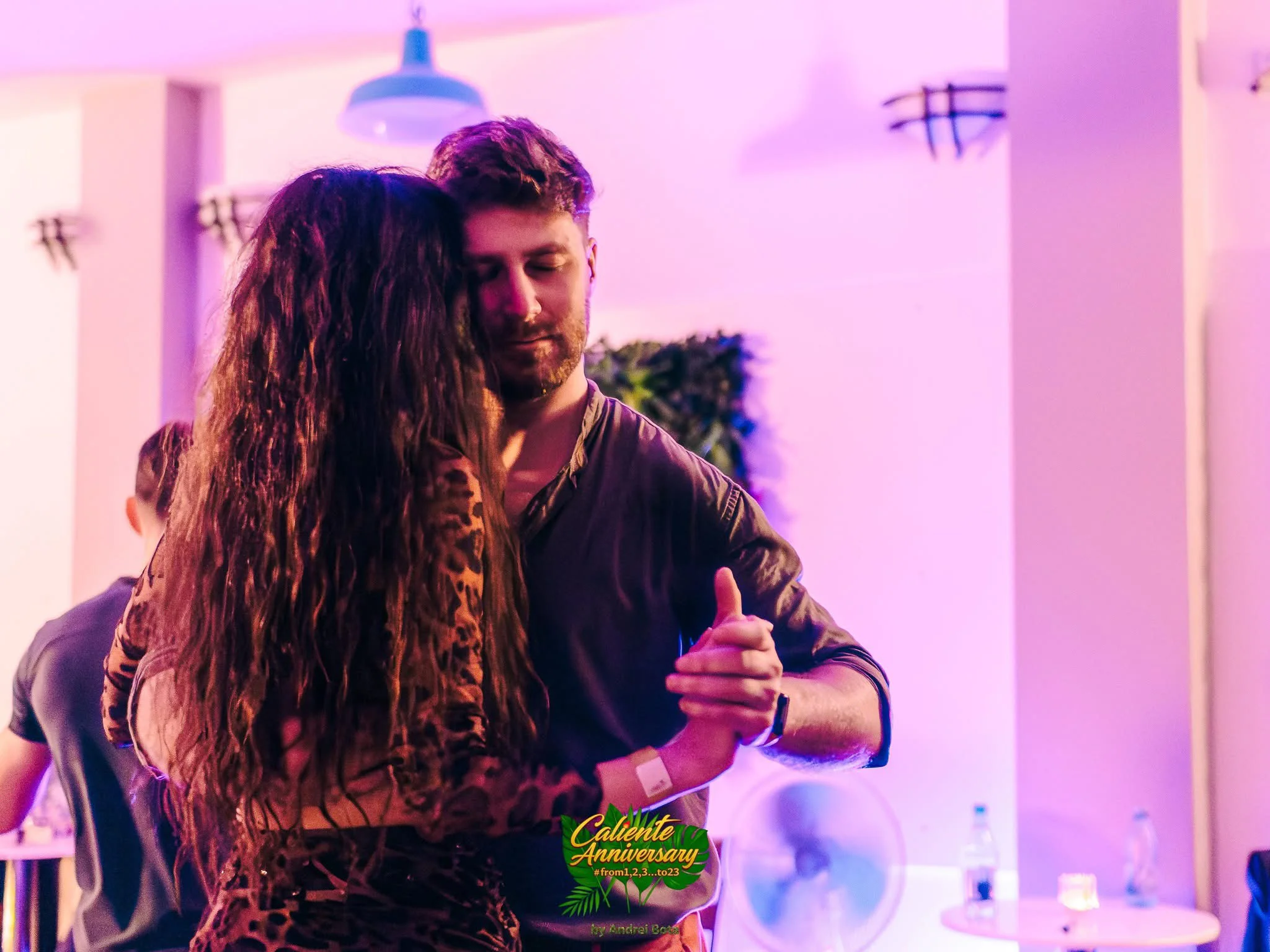 Un bărbat și o femeie dansează kizomba strâns, cu ochii închiși, în lumini roz-violet